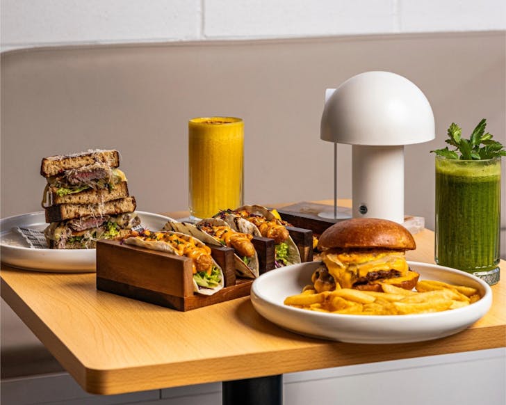 A close up image of a cafe table of food dishes and drinks including tacos, sandwiches and a burger.