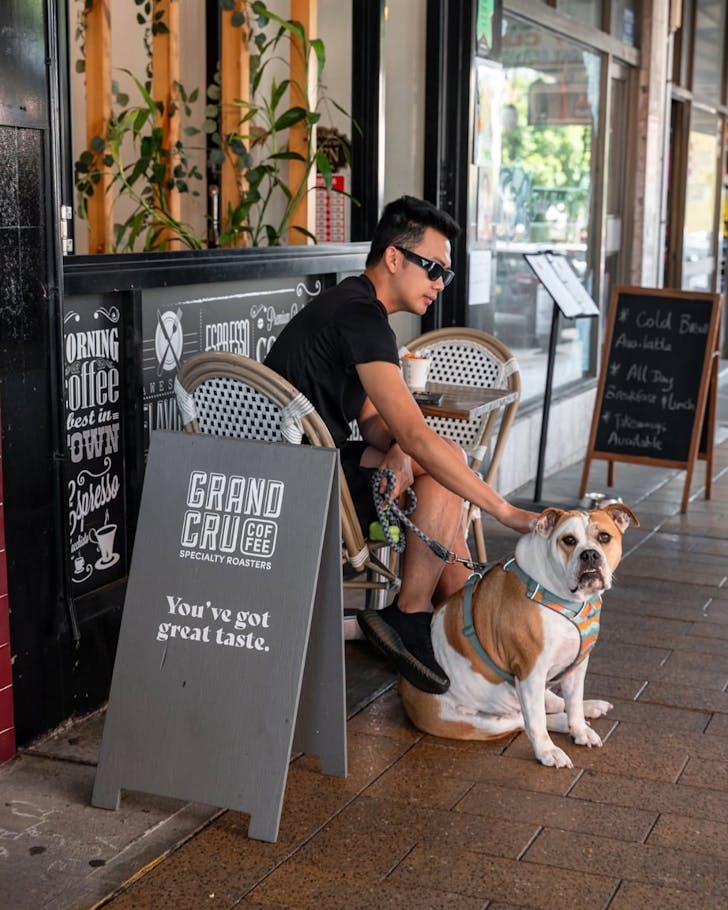 A mid-shot of a man sitting outside of Cafe The Beans petting his dog during the day time.
