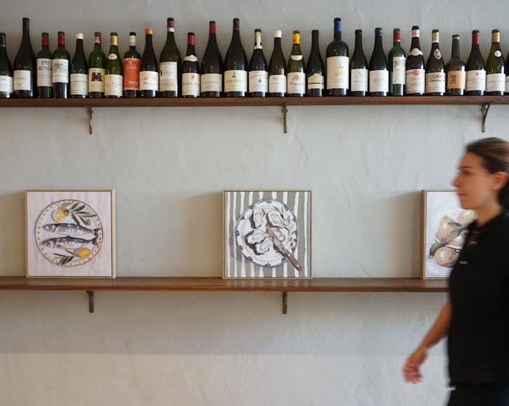 Wall at CafÃ© Margaret lined with wine bottles and still-life art, with a staff member walking pastâcapturing the relaxed Double Bay vibe.