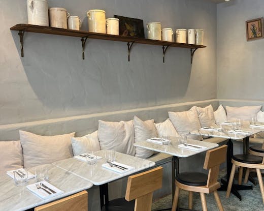 Light-filled Café Margaret dining room featuring marble tables, oak chairs and ceramic jugs displayed on a wooden shelf above linen cushions.