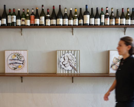 Wall at Café Margaret lined with wine bottles and still-life art, with a staff member walking past—capturing the relaxed Double Bay vibe.