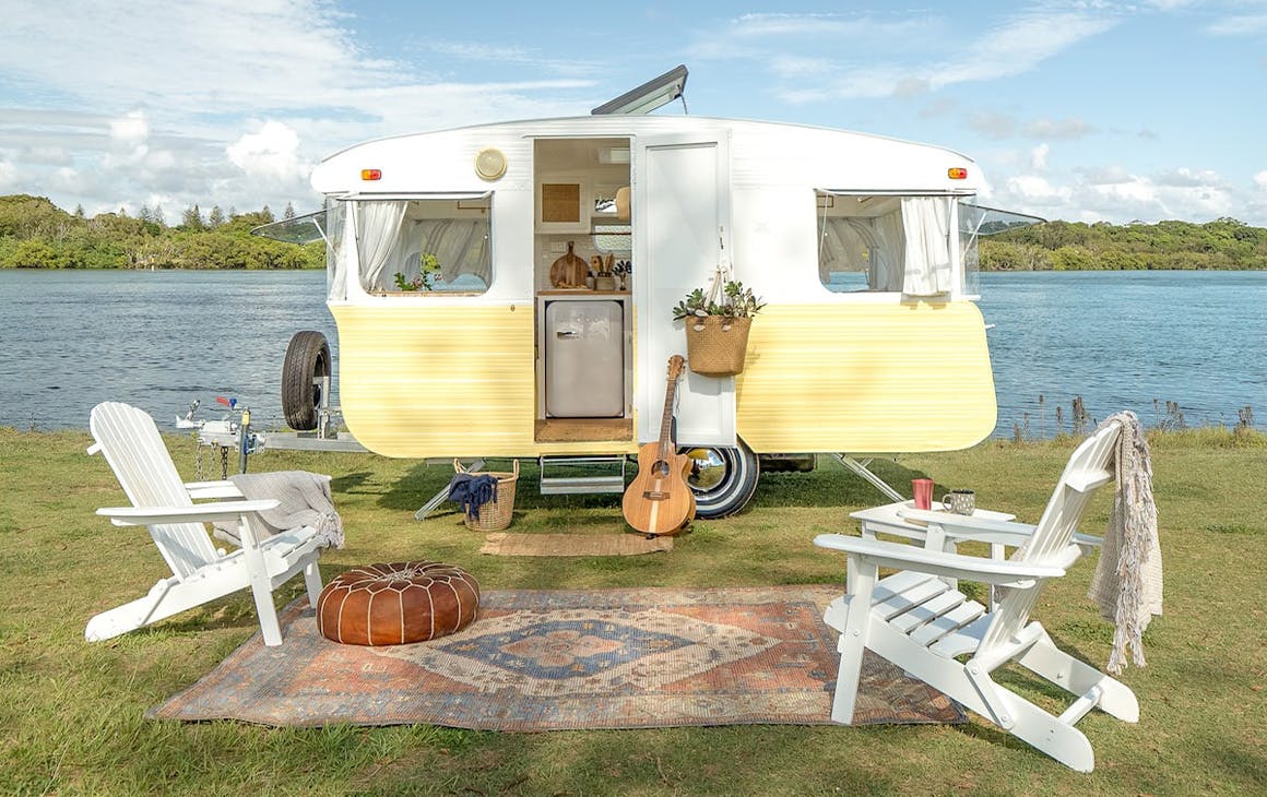 a vintage yellow caravan with white chairs out front by a river