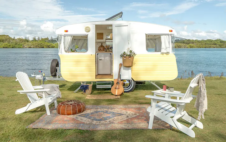a vintage yellow caravan with white chairs out front by a river