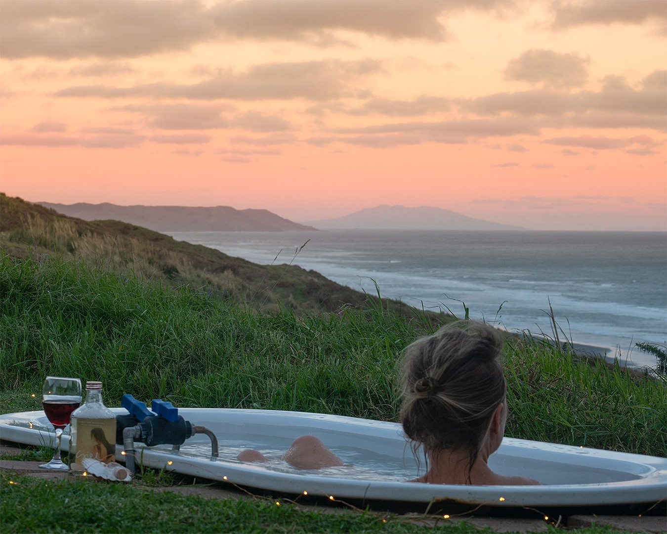 A girl looks over the sea while sitting in an outdoor bath with a glass of wine nearby. One of the best glamping experiences to be had near Auckland.