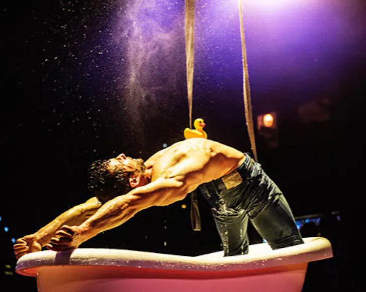 A half-naked man does a back arch over a bath tub while balancing a rubber duck on his chest. 