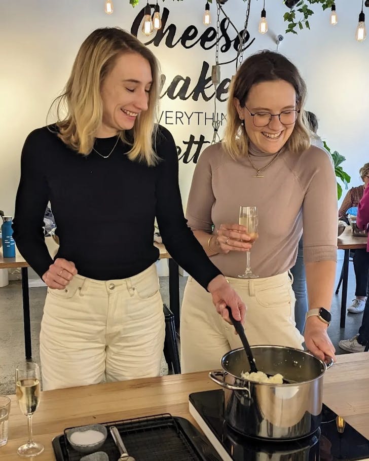 two women stir a pot of cheese while sipping champagne at a cheesemaking class
