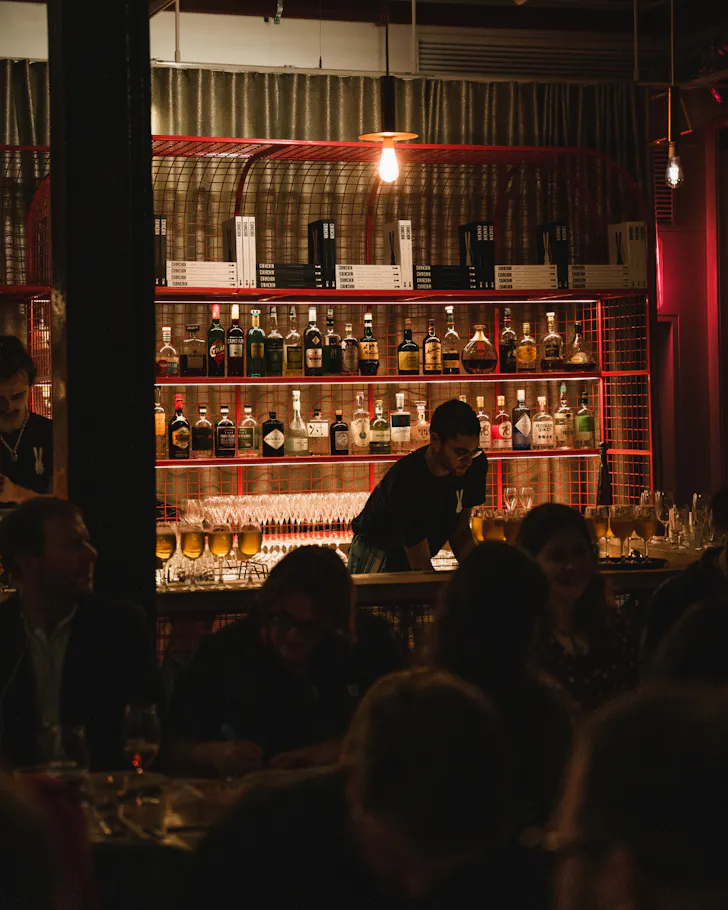 Warmly lit interior of Chin Chin restaurant, with diners filling the dining room as a bartender works beneath a backlit shelf lined with spirits and cookbooks framed by striking red metal shelving.