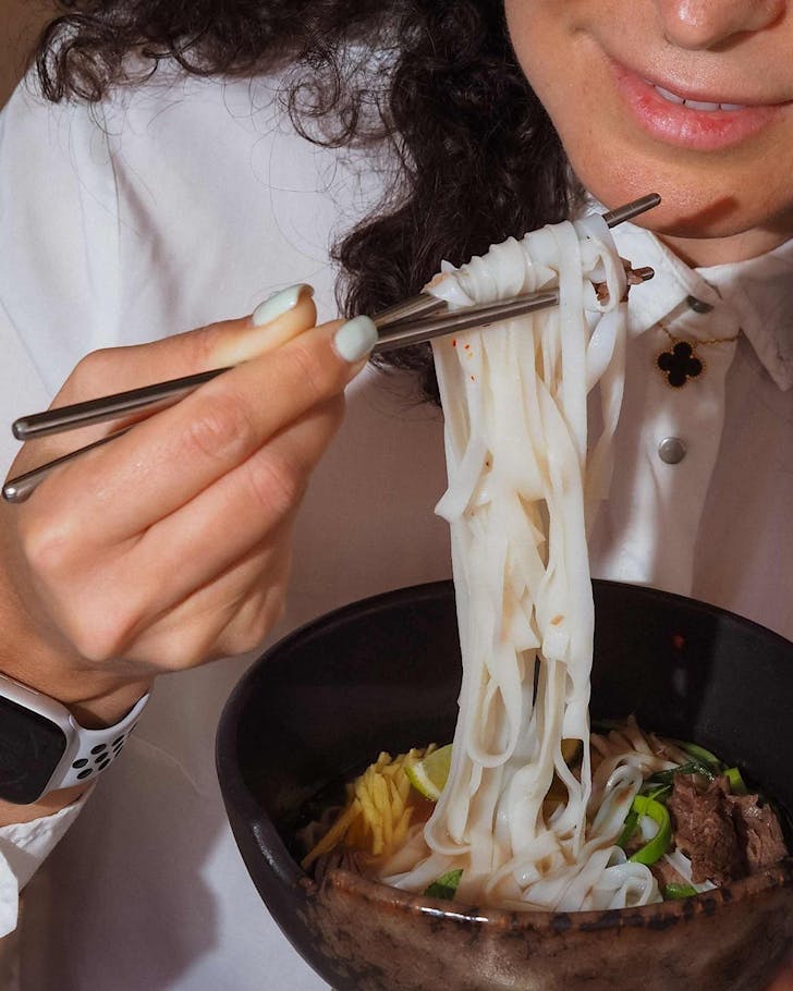 A woman uses chopsticks to pick up flat rice noodles. 
