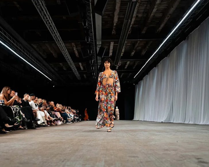 A model walks down a fashion runway wearing a bright floral two-piece outfit as a seated audience watches and photographs the show inside an industrial-style venue with exposed beams and draped white curtains.