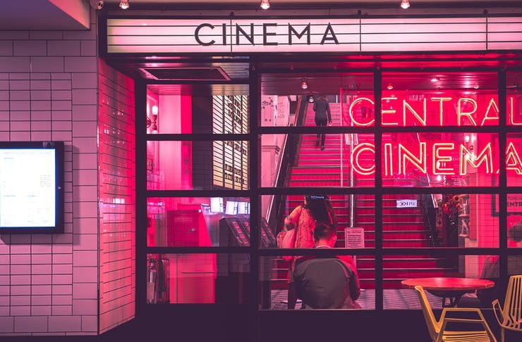A pink, neon-lit cinema shines through a glass facade. The sign above the entrance reads 'Cinema'.