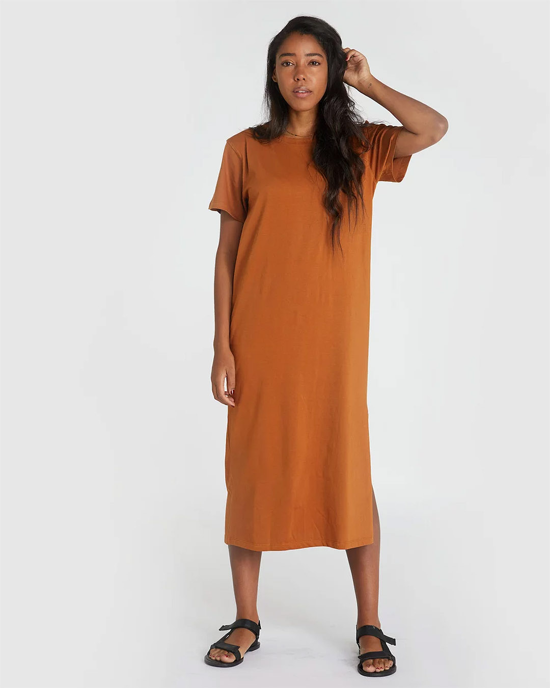A head to toe shot of a woman wearing a burnt orange mid-length t-dress, with sandals. 