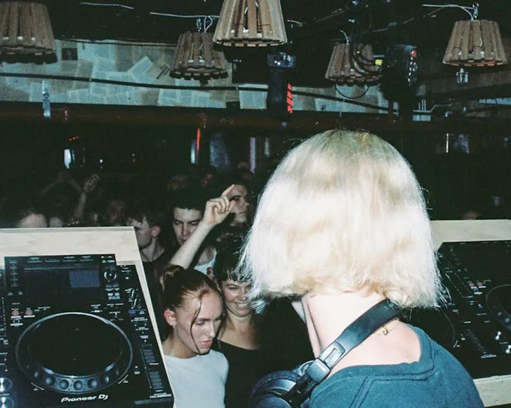 An over the shoulder image from behind a DJ as she faces a crowd of people dancing to her music.