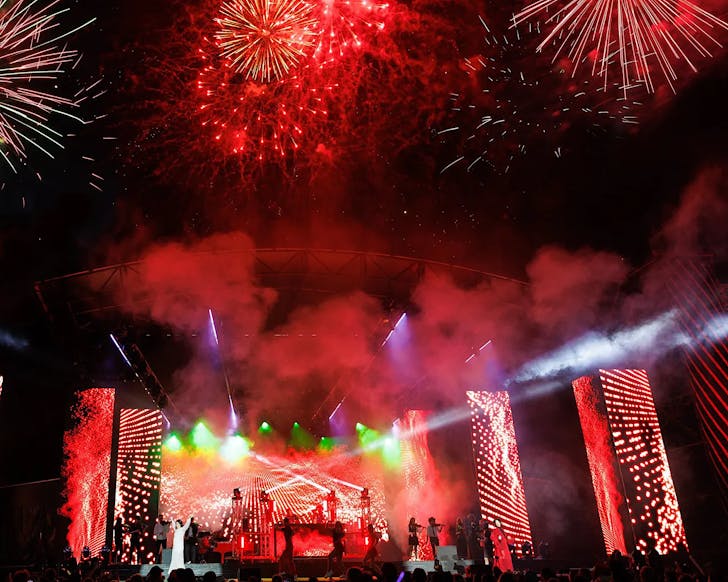 A singer dressed in white with their arms up as the night sky above them explodes with red fireworks.