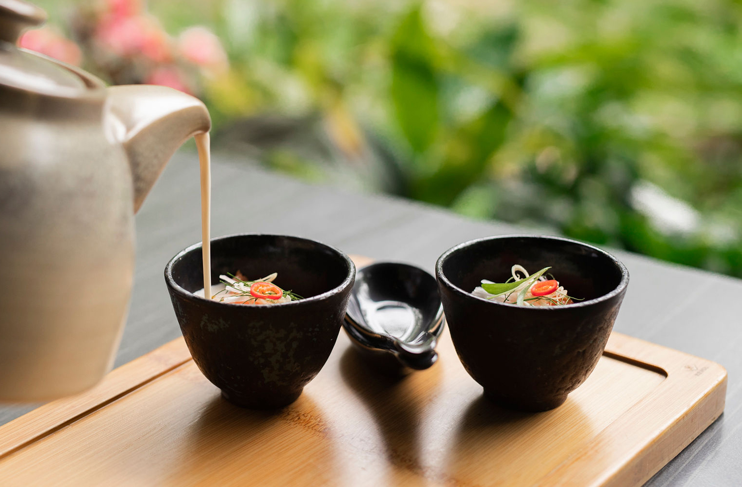 A top lay image of a teapot pouring coconut salmon soup into two small black bowls. 