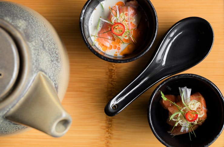 A flatlay image of a teapot, two black bowls filled with Spirit House's salmon coconut soup and a black soup spoon. 