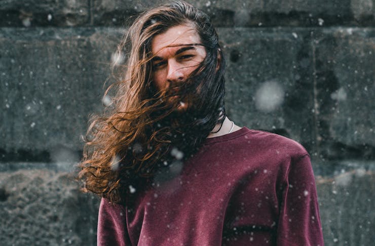 A man with long hair blowing in the wind, stands outside while light snow falls.