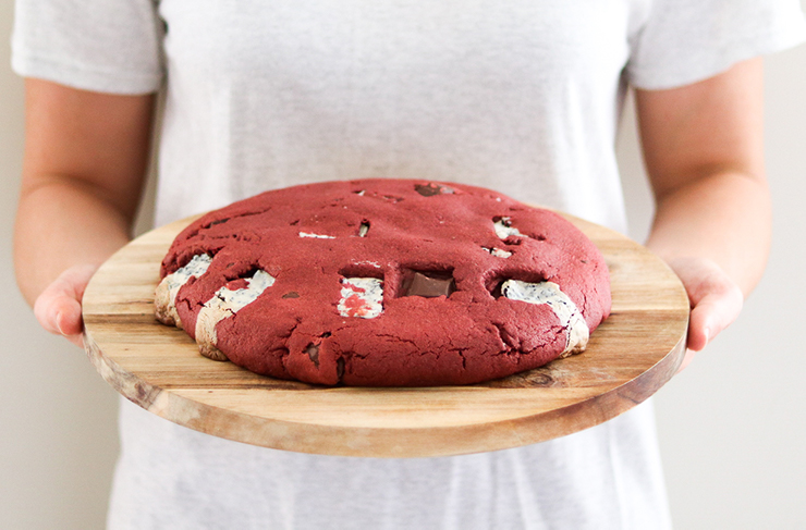 A person in a white shirt holding a wooden board atop which sits a large red velvet pizza cookie.
