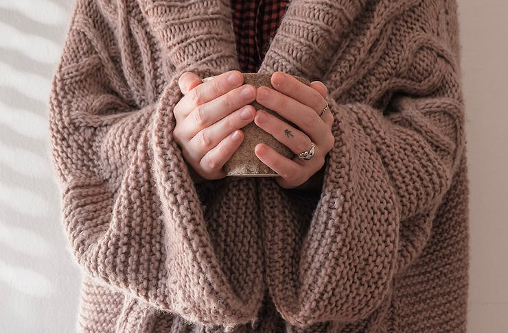 A woman in an oversized brown knit sweater stands with her hands on a hot drink.