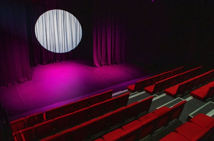 The inside of Covert Theatre—classic amphitheatre style red velvet setting and purple lighted stage, big silver spotlight on the backdrop between velvet curtains.