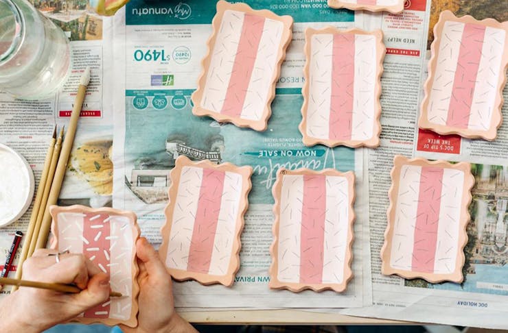 6 small plates shaped like iced vovo biscuits on a table covered in newspaper, being painted by a hand