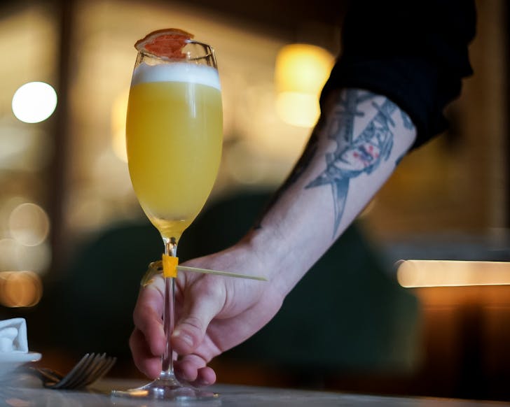 The tattooed arm of a Bossi server places down their Daffodil Day cocktail on the table.