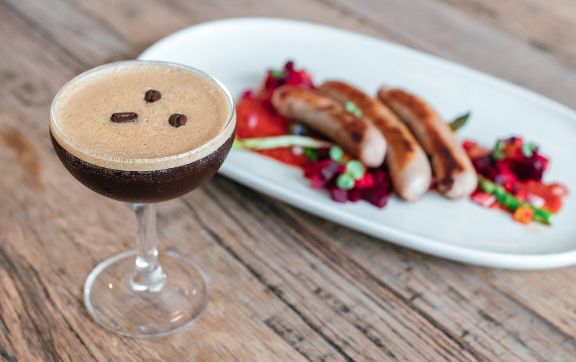 an espresso martini in front of a plate of sausages