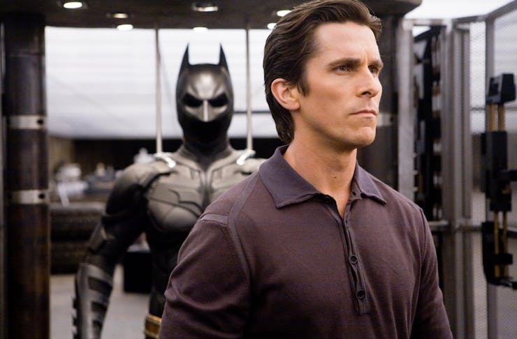 Christian Bale as Bruce Wayne, standing with his back turned to his Batsuit.
