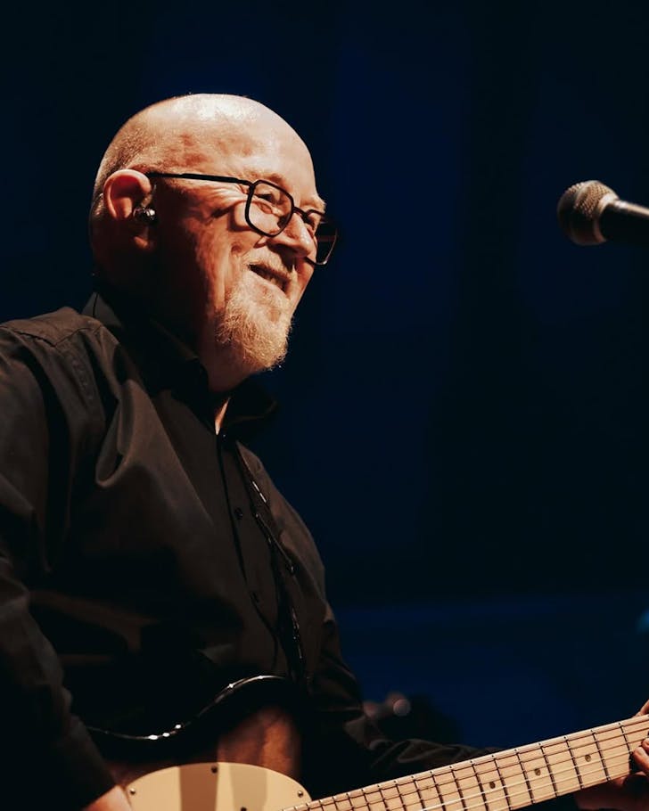 Close-up of a spotlighted Sir Dave Dobbyn holding his guitar, performing on a dark stage.