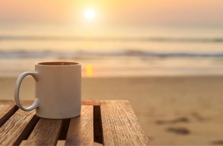 A white mug on a bench at the beach at sunrise.