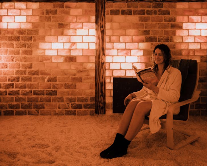 A lady sitting on a chair in a salt room. The room looks normal except for red lighting and a floor that is covered in a thick layer of salt. The lady looks relaxed as she sits, smiling and reading.