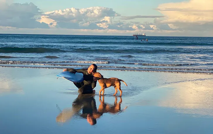 Woman holds surfboard at the beach with her dog