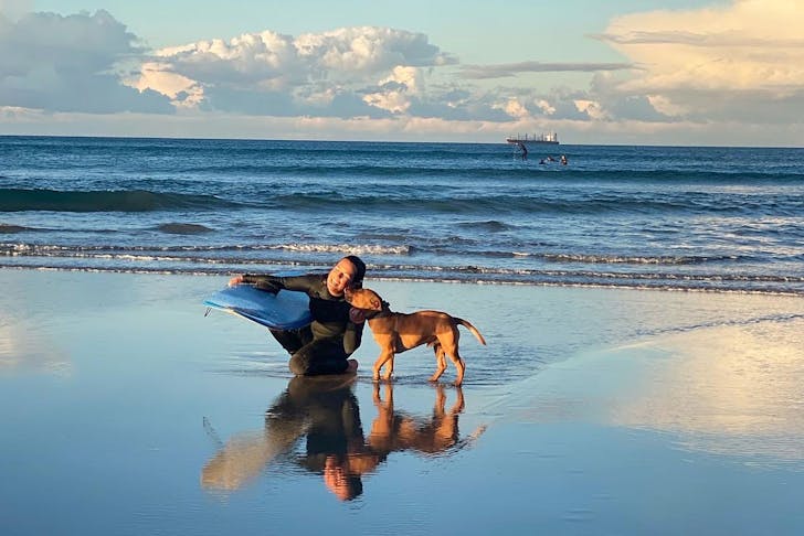 Woman holds surfboard at the beach with her dog
