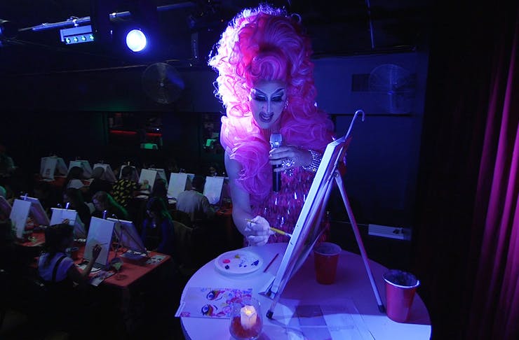 A drag queen paints away at Dragvine, close up shows her canvas, hand and a bit of her laughing face.