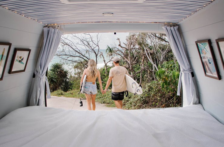 A photo taken from inside a camper van, looking out at a couple walking towards the beach with surfboards.