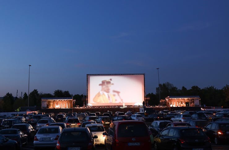 Rows of cars sit in front of a large outdoor movie screen at a drive-in cinema.
