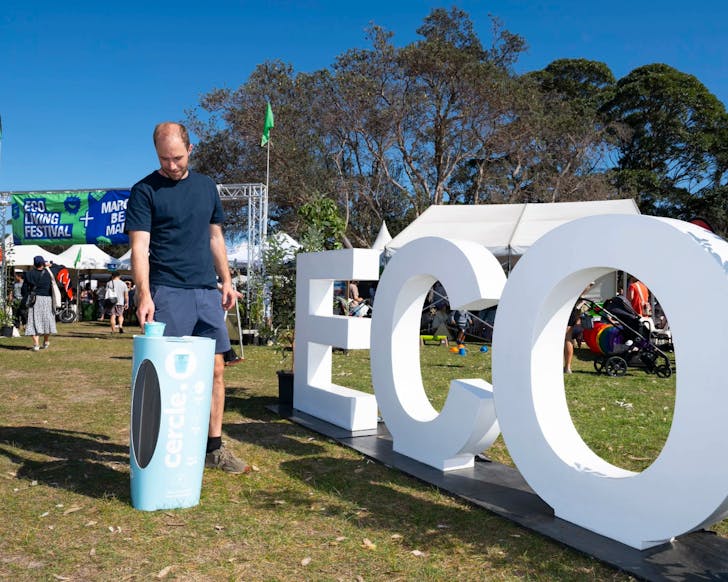 An image of a man in an outdoor venue standing next to a giant sign that reads 'ECO', and he is filling up his water at a recycled water station.