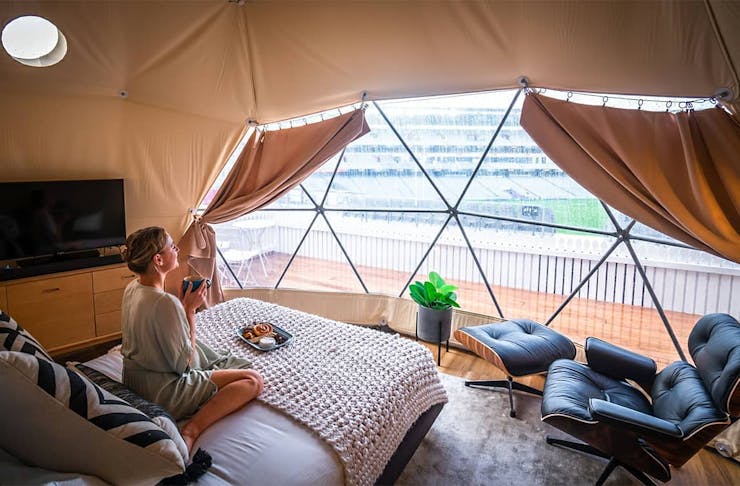 A girl sits on the bed in the Eden Park glamping accommodation looking pleased as punch with a cup of tea in her hands.