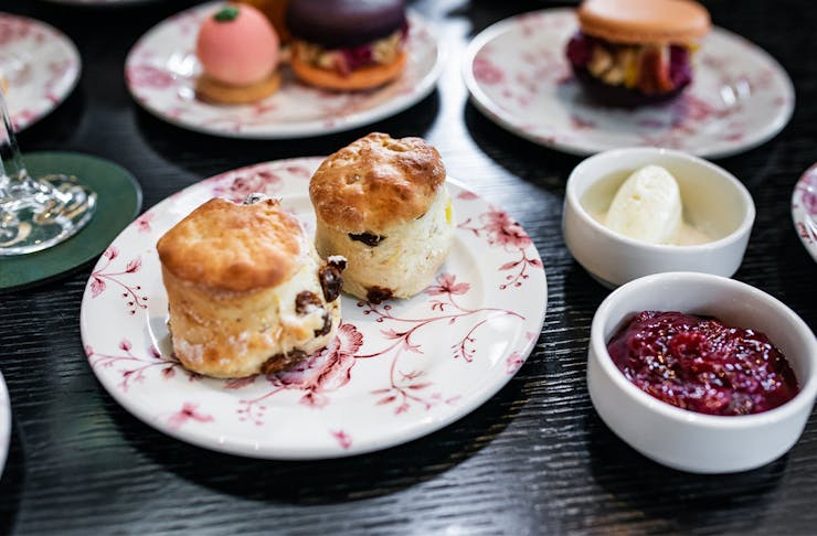 two scones on a floral plate, with jam and cream in seperate bowls next to the plate and other high tea treats in background