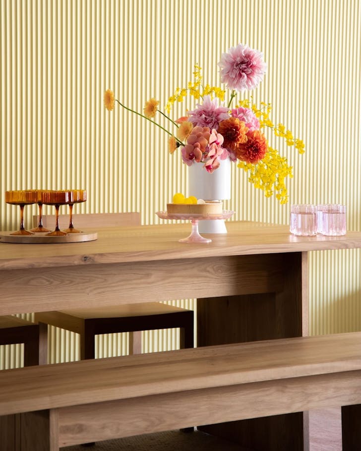 A minimal wooden dining table topped with pastel glassware and an bunch of flowers in a clear vase.