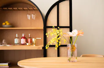 A wooden table topped with a vase of flowers in front of a rattan bar cabinet by RJ Living.