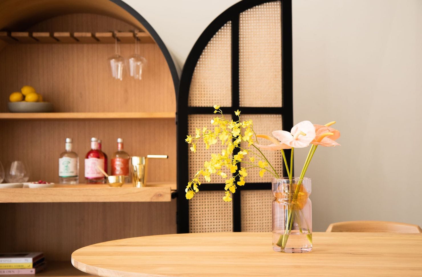 A wooden table topped with a vase of flowers in front of a rattan bar cabinet by RJ Living.