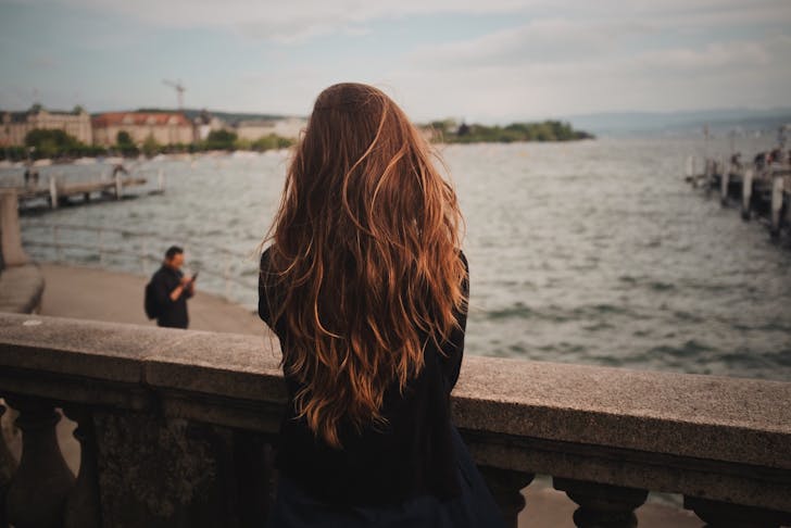 A girl with long wavy brown hair standing over a stone balcony, looking out at the sea.