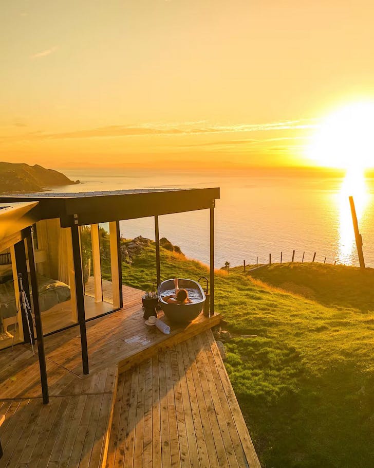 Someone enjoys a sunset soak in a freestanding, outdoor bath before an expansive coast.