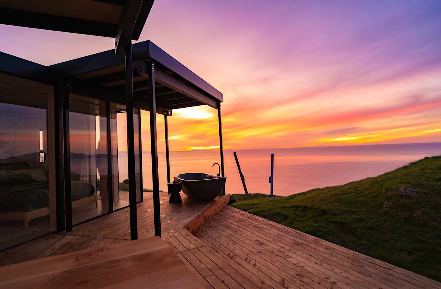 A vibrant purple and orange coastal sunset behind a modern Airbnb with an outdoor deck area and freestanding outdoor bath.