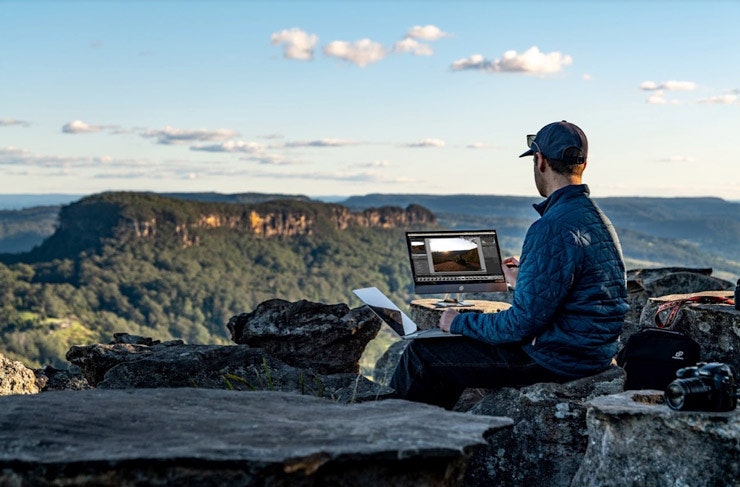 A person with a computer monitor sitting on a rocky cliff near the Blue Mountains.