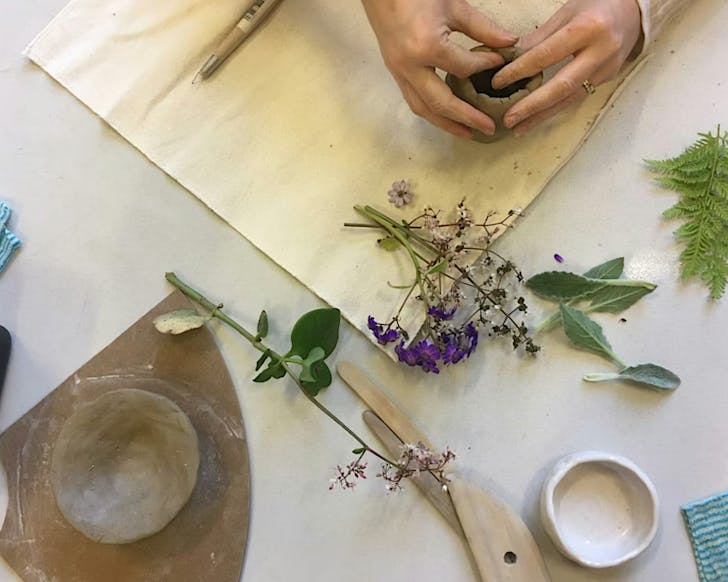 Someone works on soft clay during a pottery workshop, while other instruments and some purple flowers strew the table.
