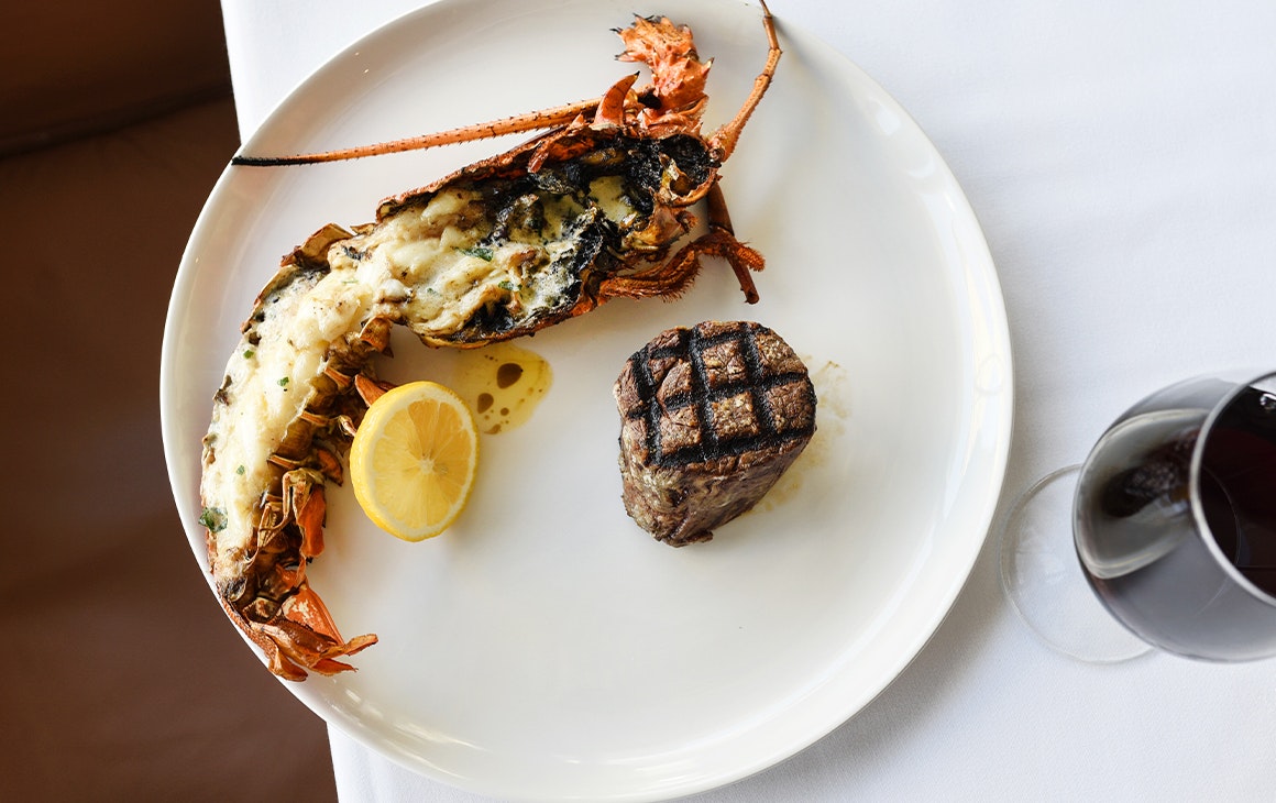 a steak and a half rock lobster on a plate with a glass of wine