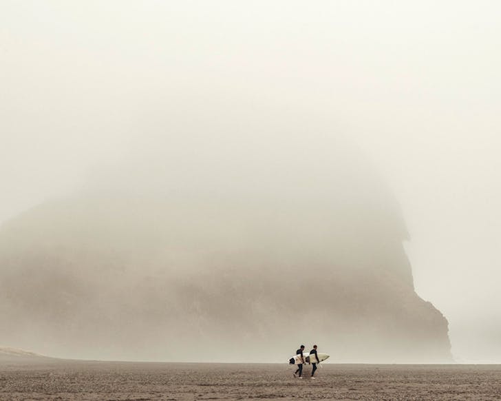 Two surfers carry their boards, walking before a mist-shrouded Lion Rock at Piha Beach.