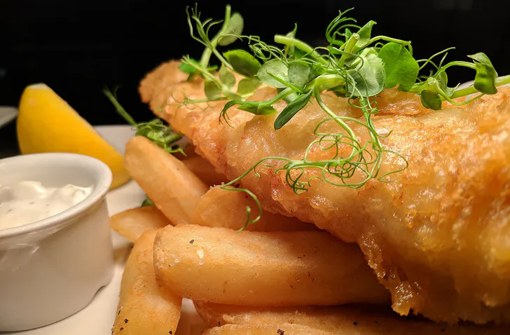 Fish and chips sit on a plate with a wedge of lemon and tartare sauce on the side.