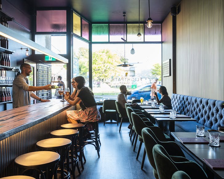 An interior image of Flight Path Wine Bar with people smiling and sitting at the bar and table while coloured light filters in through the stained glass window at the entry.